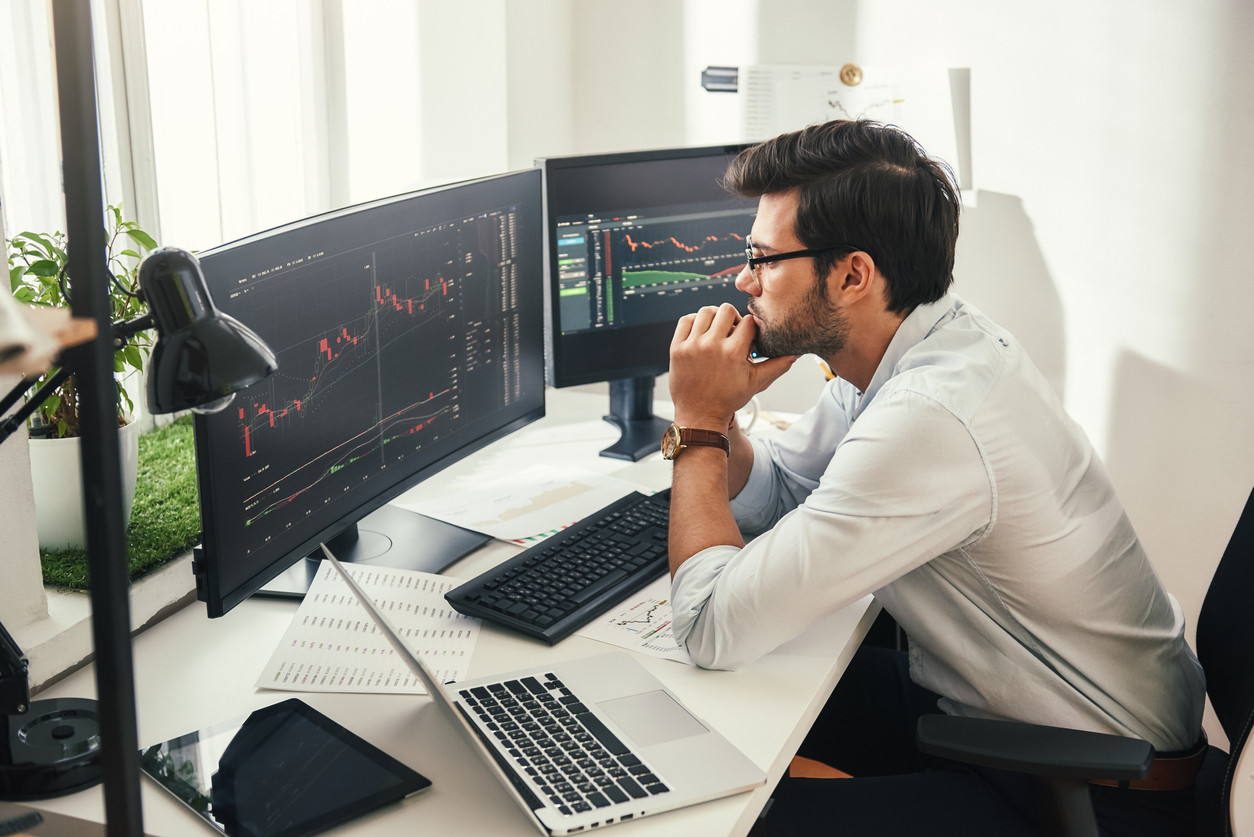 Successful trader. Back view of bearded stock market broker in eyeglasses analyzing data and graphs on multiple computer screens while sitting in modern office.